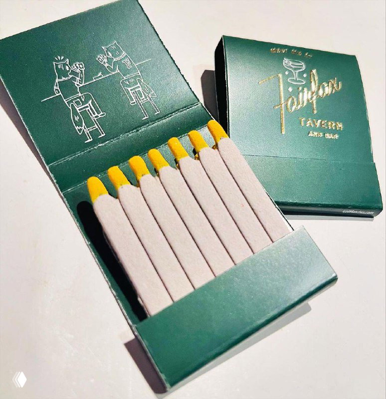 Тёмно‑зелёная коробка с золотистым тиснением и иллюстрацией, внутри спички с жёлтыми головками; упаковка выглядит как брендированный сувенир из кафе или лавки.