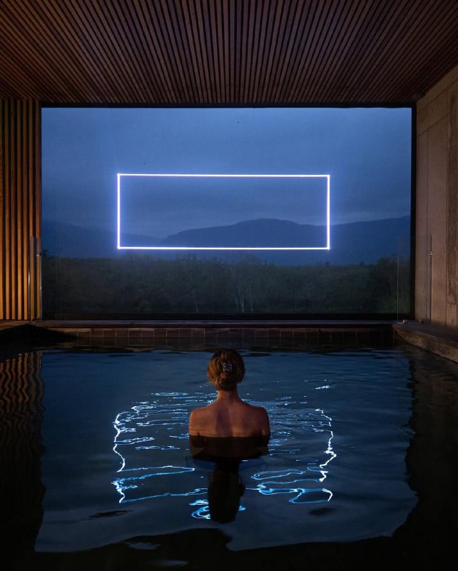 Человек в воде смотрит на прямоугольную неоновую рамку над ландшафтом — кадр, объединяющий воду, свет и окружающий пейзаж в инсталляции.