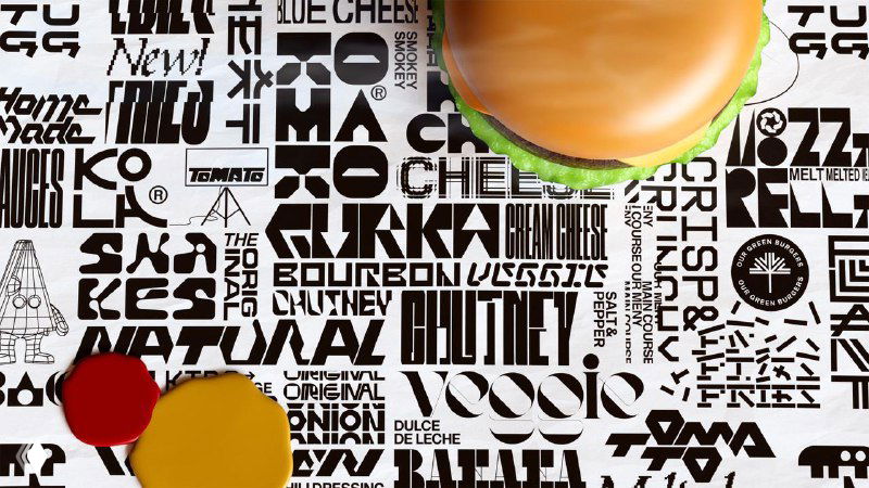 Крупный план рабочего стола с коллажем из типографики, логотипов и фонов, на которых стоит часть гамбургера — дизайн упаковки и паттернов.