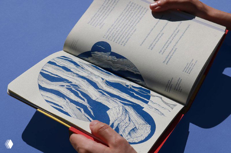 Крупный план рук, переворачивающих страницы книги; видны вырезанные графические формы, насыщенные синие иллюстрации и фактура бумаги с тенью от рук.