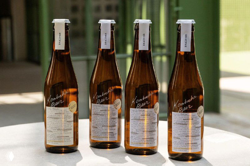 Четыре бутылки Brain+er выстроены в ряд у окна: прозрачное стекло, текстовые этикетки и белый леттеринг заметны на солнце, съёмка подчёркивает фактуру упаковки.