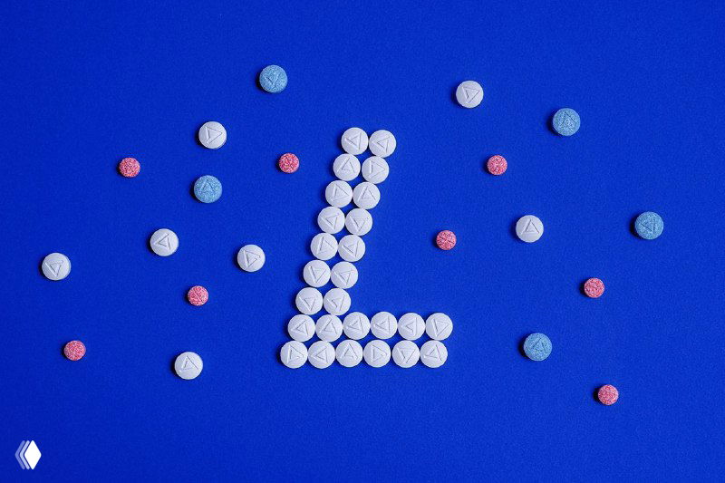 Таблетки, разложенные на синем фоне в форме буквы L рядом с рассыпанными разноцветными пилюлями — стилизованная композиция для упаковки Nastizol.