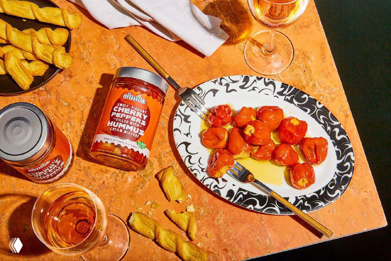 Тарелка с веганскими антипасти и банки Almito на столе с закусками и бокалами — тёплая съёмка, показаны цвета этикеток, подача.