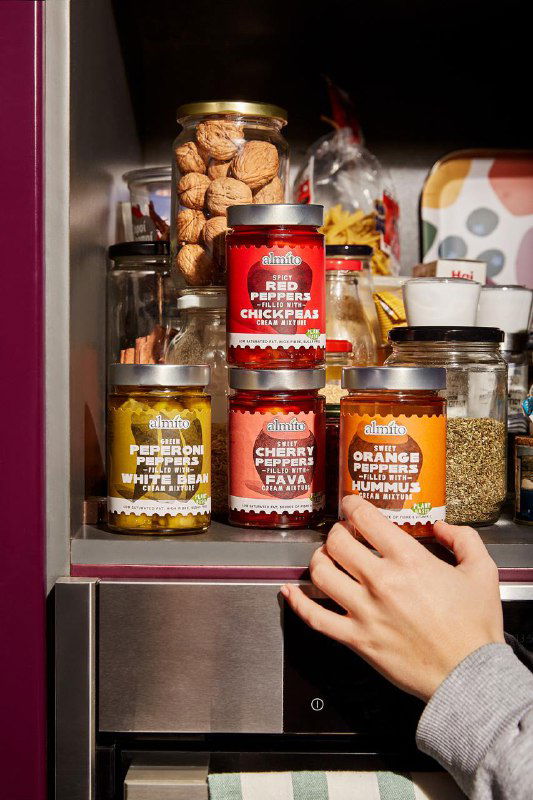 Несколько банок Almito разных вкусов на полке-шкафу, одна рука берёт банку; видно разнообразие этикеток и размеры упаковок.