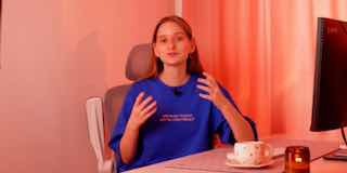 Кадр документального ролика: ведущая в синей футболке сидит за рабочим столом, активно жестикулирует; на столе чашка и свеча, фон розового тона