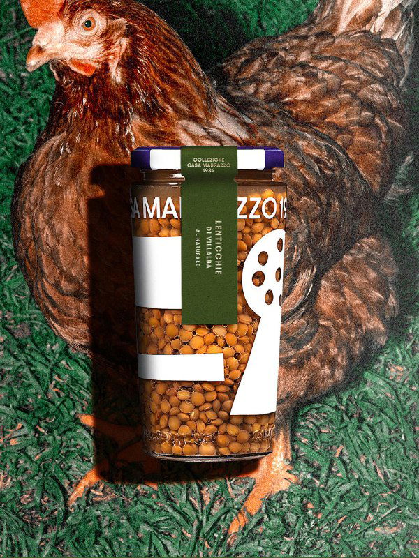 Одна банка Casa Marrazzo 1934 лежит на траве рядом с курицей; этикетка с крупной иллюстрацией и текстурой продукта.