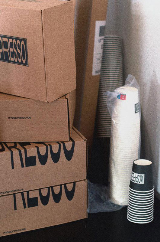 Стопка коробок и бумажных стаканчиков рядом с упаковкой Mospresso; композиция подчёркивает брутальную простоту брендинга, логотип и упаковочные материалы.