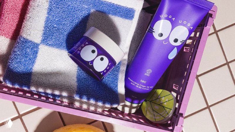 Тюбик и баночка Uoga Uoga на плетёной поверхности с полотенцем и фруктом — атмосферная продуктовая съёмка с акцентом на упаковку.