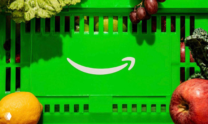Зелёная пластина или ящик с белой стрелкой‑логотипом Amazon среди овощей на фоне — визуальная отсылка к Amazon Fresh и упаковке для продуктов.