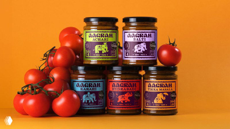 Несколько баночек соусов Aagrah на оранжевом фоне рядом с помидорами; контрастные яркие этикетки с орнаментами и иллюстрациями слонов.