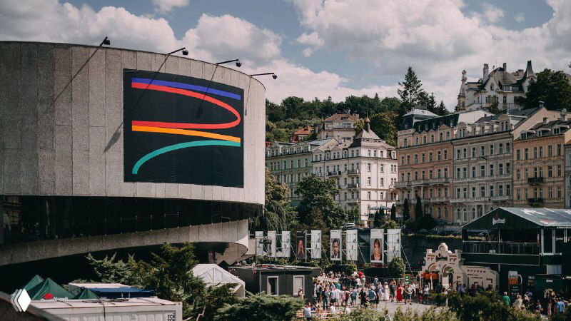 Городской кадр: здание в Карловых Варах с большим световым экраном, на котором отображены цветные дуги айдентики 57-го кинофестиваля KVIFF, люди и городской пейзаж.