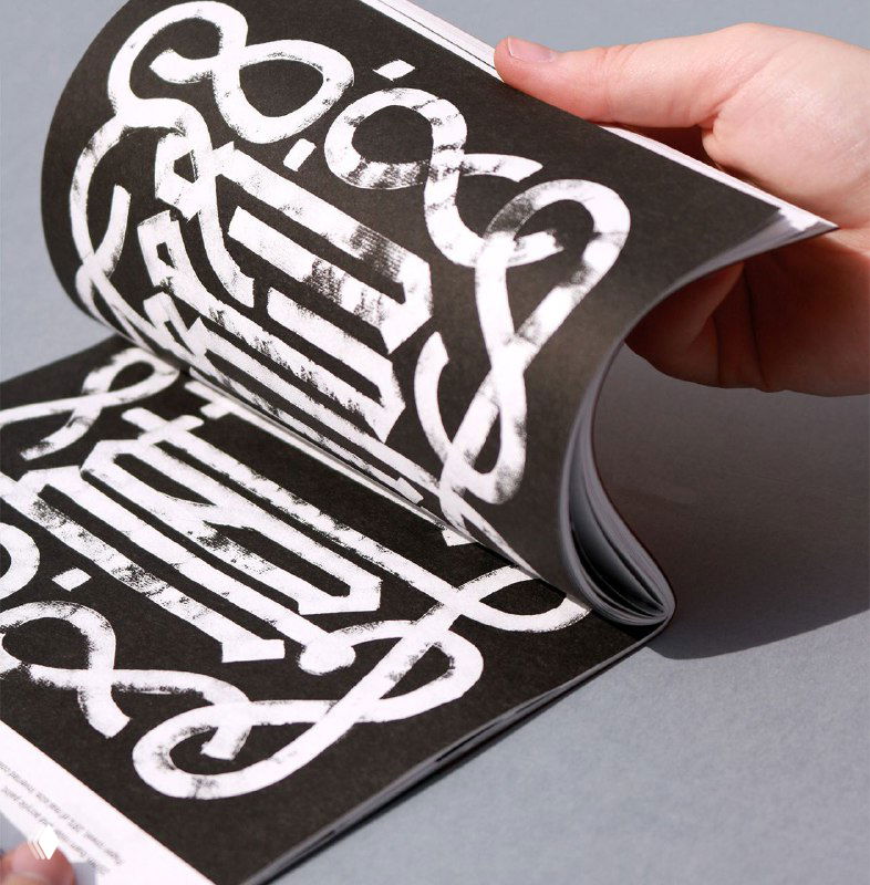 Крупный план внутренней страницы зина «Blackletter. Textura» с белой мастикой на чёрном фоне и каллиграфическими элементами в руке держателя.