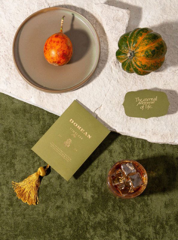 Плоская композиция: визитка Dorean с кисточкой, бокал со льдом и декоративные фрукты на текстурной поверхности — стилистика и материалы айдентики.