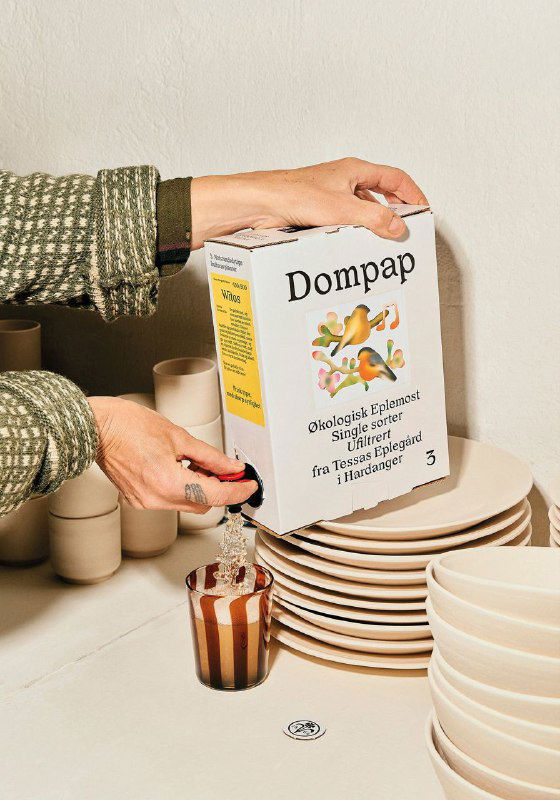 Человек держит упаковку Tessas Eplegard над тарелками; видно название Dompap и иллюстрацию, подчёркивающую напиток и эстетику упаковки.