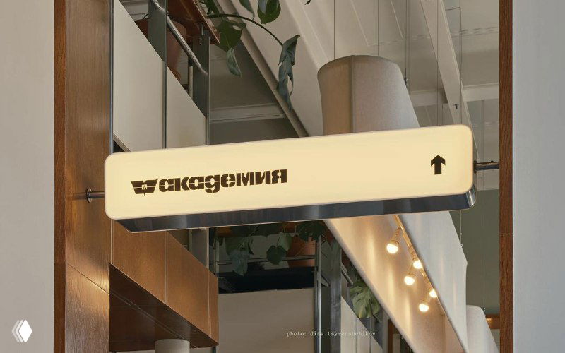Интерьерный указатель с надписью «академия» на светлом прямоугольном табло, стрелка направления и часть внутреннего пространства магазина‑кафе.