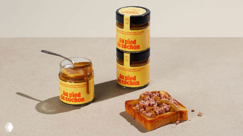 Несколько стеклянных банок с этикетками Au Pied de Cochon разного размера на столе рядом с кусочком хлеба с паштетом; минималистичная презентация.