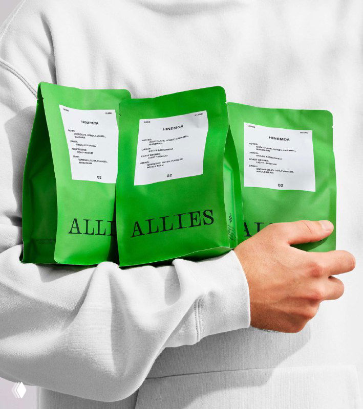 Человек держит несколько зелёных пакетиков кофе Allies у груди — акцент на материальности упаковки и фирменном зеленом цвете.