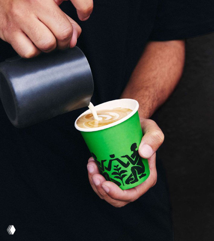 Бариста наливает молоко в чашку, держа зелёный стакан Allies — демонстрация брендированной подачи кофе в реальной сцене.