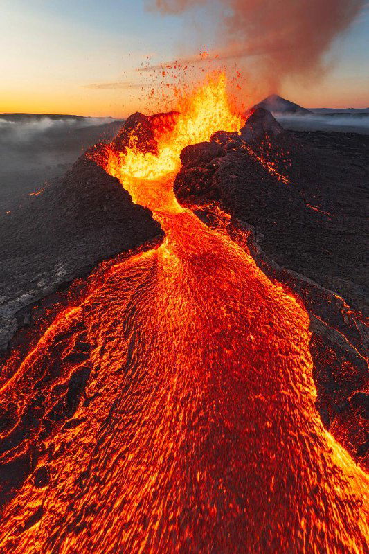 Активный вулкан с лавовым потоком: яркие оранжево-красные потоки лавы контрастируют с тёмной поверхностью окрестностей, снято сверху.