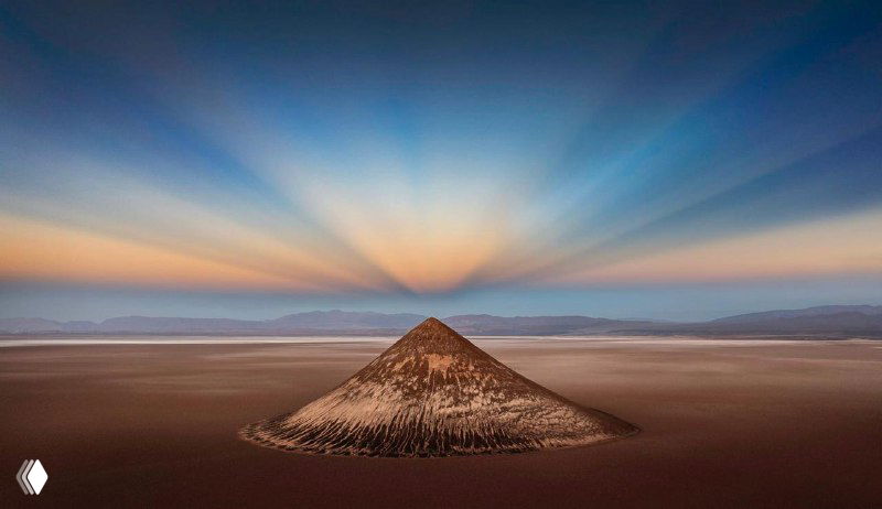 Симметричный пейзаж с коническим холмом и лучами света на рассвете: аэрофотография выделяет форму, перспективу и тональные переходы.