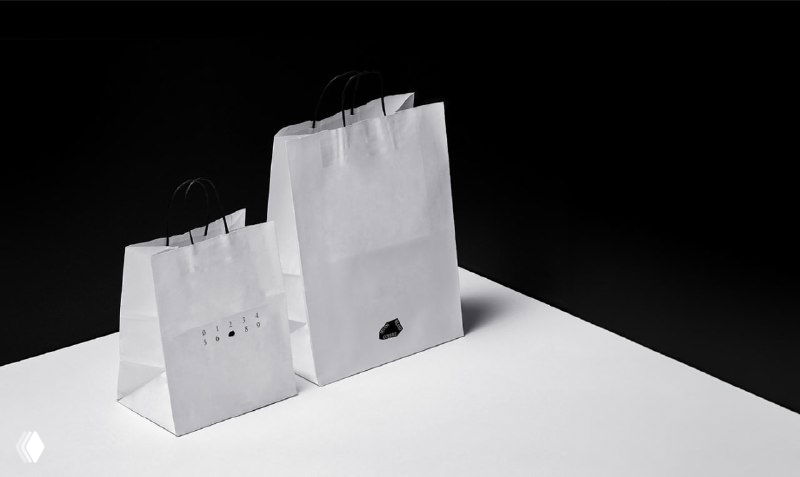 Фотография белых бумажных пакетов с небольшим чёрным знаком логотипа в минималистичной студийной композиции на светлом фоне.