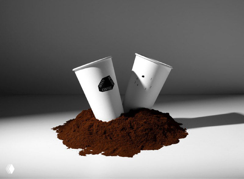 Две белые одноразовые чашки с чёрным семиугольным знаком, расположенные на горке молотого кофе в художественной посадке освещения.