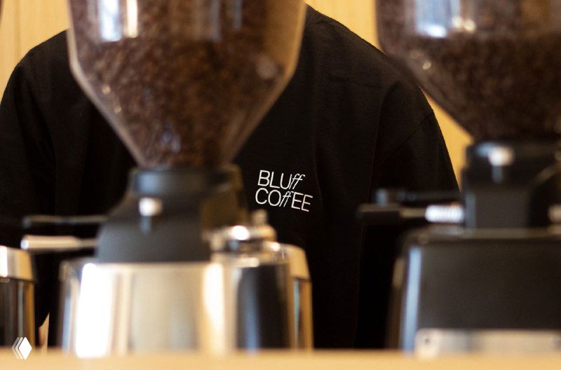 Крупный план кофемолок на барной стойке; на одежде бариста видно шитое лого «BLUff COFFEE», иллюстрирующее применение айдентики в мерче.