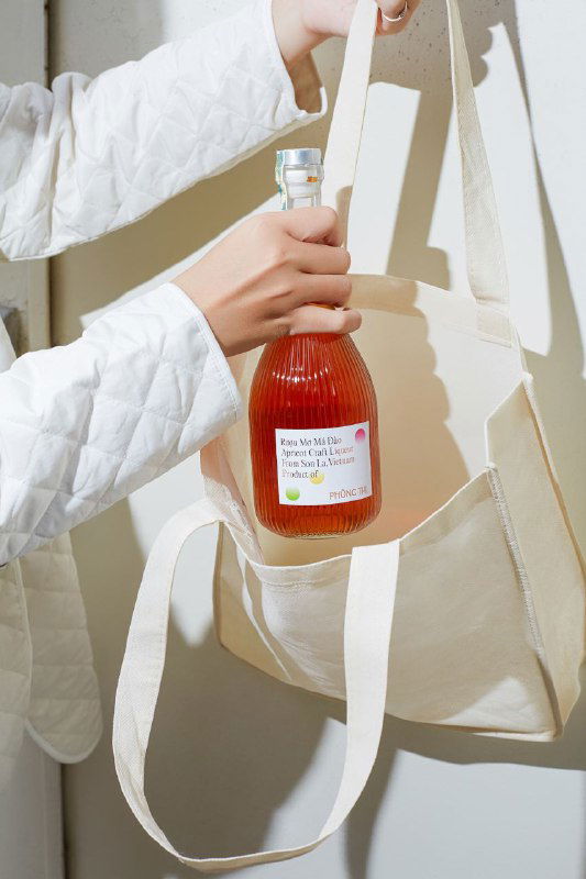 Рука держит рифлёную бутылку Phung Thi в текстильной сумке на нейтральном фоне — демонстрация портативности и эстетики бутылки при повседневном использовании.