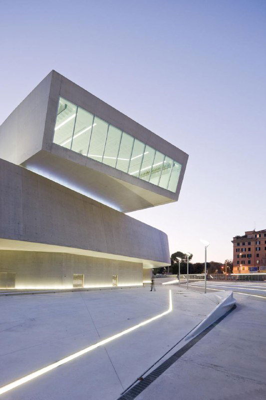 Контрастная современная пристройка с выступающим стеклянным объёмом и чистыми бетонными поверхностями — минималистский музейный фасад в вечернем свете.