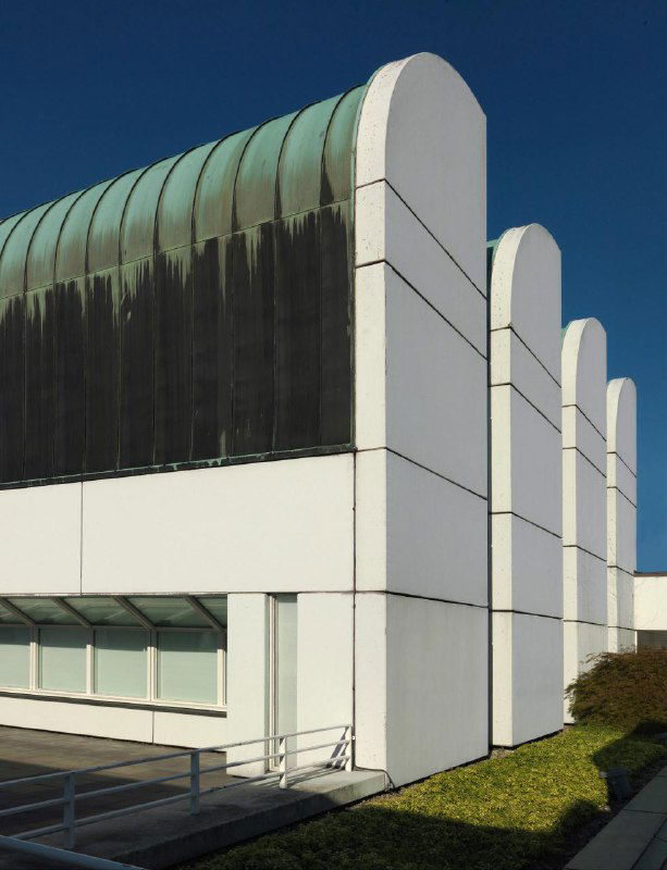 Белые округлые пилоны и зелёная кровля на фоне неба; вид сбоку на музейный корпус с плавными формами и выразительной структурой фасада.