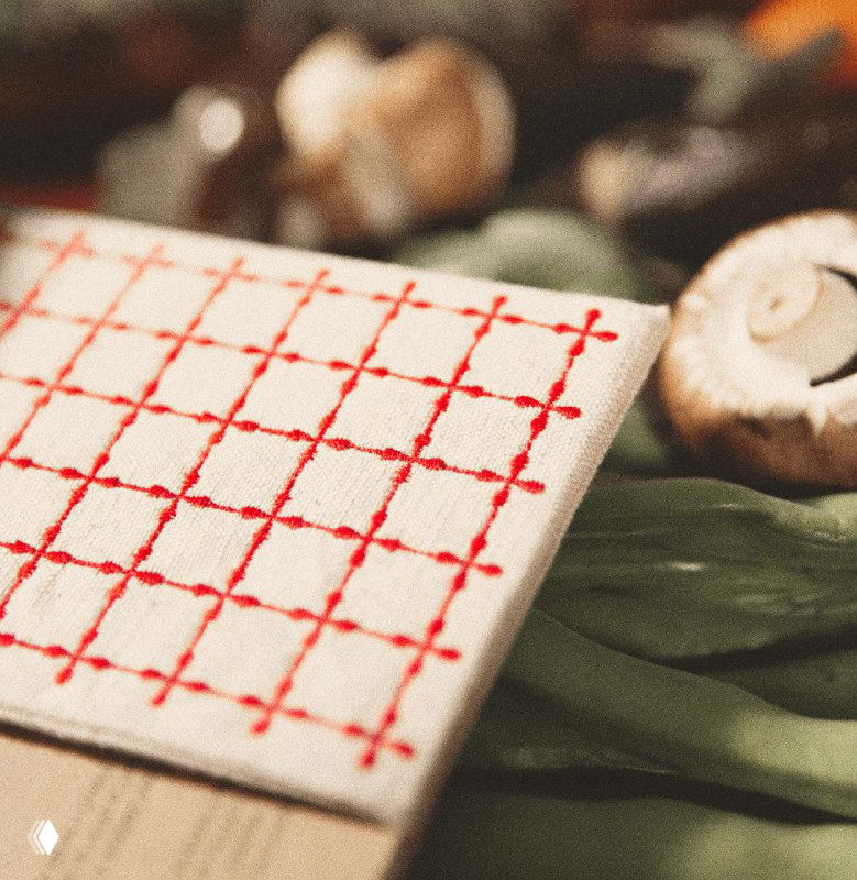 Деталь вышивки на тряпичной обложке в виде красной сетки на фоне грибов и зелёных листьев; акцент на ручной стилизации.