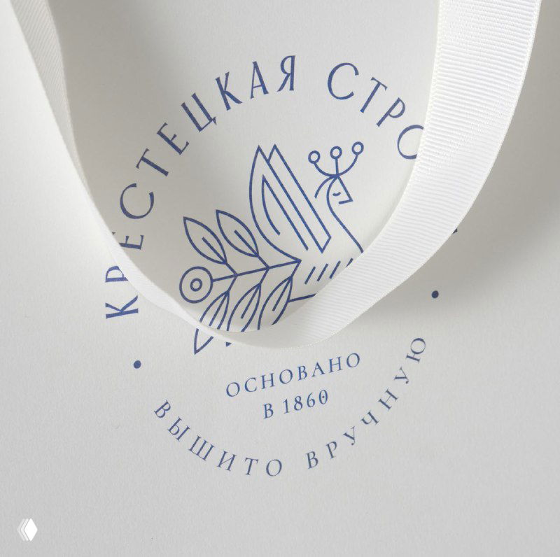 Графический знак бренда, отпечатанный синим на бумажной ленте сумки: стилизованная веточка, орнамент и надпись «Крестецкая строчка».