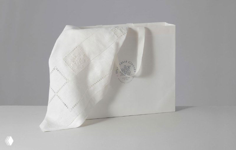 Товарный пакет и льняная скатерть с тонкой ручной вышивкой: минималистичная съемка продукта, показывающая фактуру ткани и фирменный знак бренда.