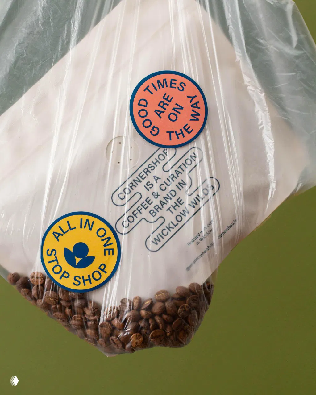 Прозрачный пакет с кофейными зёрнами и круглыми наклейками «Good times are on the way» и «All in one stop shop», элементы айдентики видны крупно.