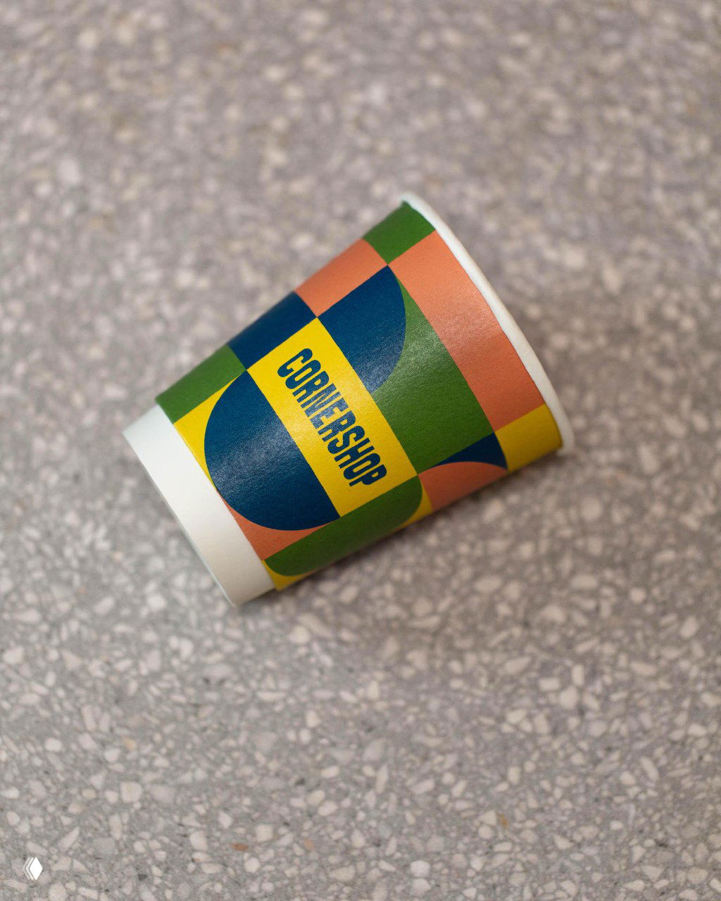 Близкий предметный кадр бумажного стаканчика с яркой геометрической печатью и надписью CORNERSHOP, деталь фирменного узора на фактурной поверхности.