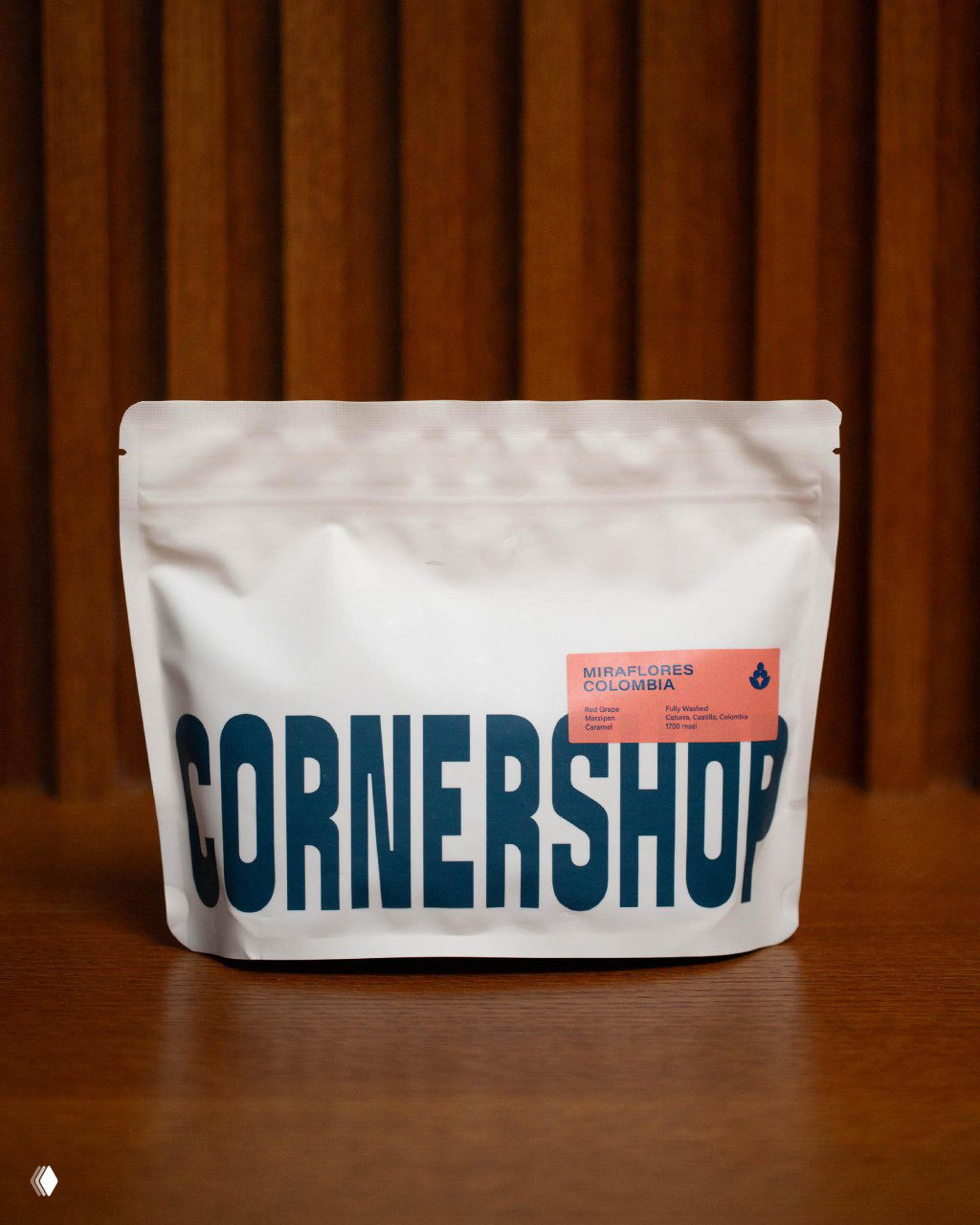 Пакет кофе CORNERSHOP на деревянной плоскости с крупным синим логотипом и небольшими этикетками Miraflores Colombia, стильная студийная съёмка.