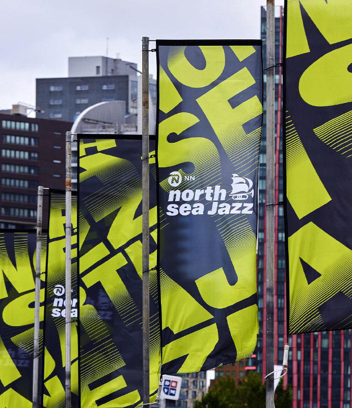 Флаги с жёлто‑зелёной кинетической типографикой North Sea Jazz, развевающиеся на улице — пример брендирования городской среды и навигации фестиваля.