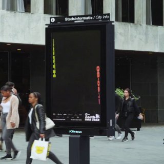 Уличный билборд с чёрным фоном и вертикальной кинетической типографикой North Sea Jazz, вид в городской обстановке с пешеходами.