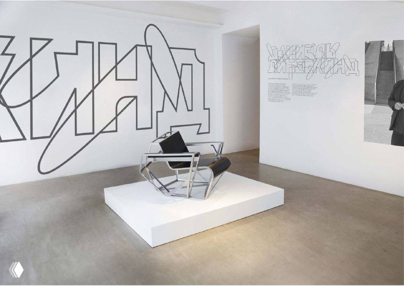 Белое минималистичное помещение с крупной типографической росписью на стене и простым подиумом с креслом, экспозиция в галерейном пространстве