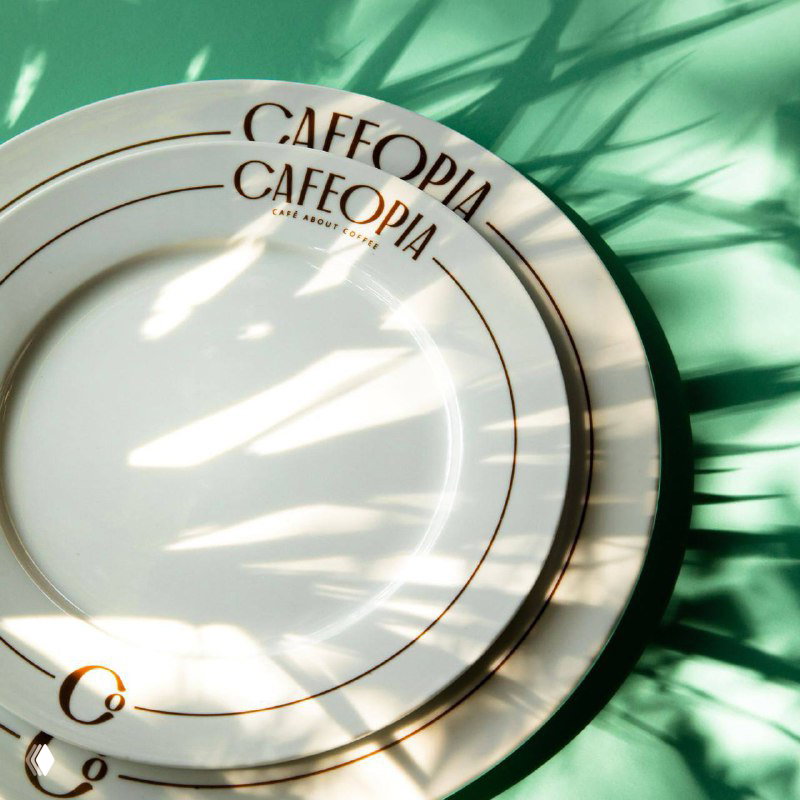 Тарелки Cafeopia с тонким золотым шрифтом логотипа на белой керамике: свет и тени создают атмосферу элегантности и ар‑деко настроения.