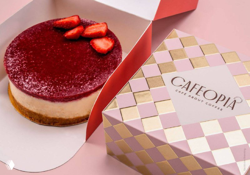 Коробка десерта Cafeopia с клетчатым орнаментом в пастельных тонах и открытым чизкейком внутри — сочетание упаковки и продукта в кадре.