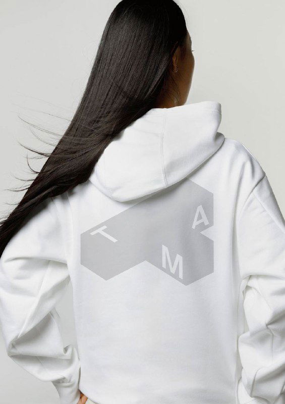 Белая худи, сфотографированная со спины, на которой крупным планом размещён серый трёхмерный логотип TMA — пример брендированной одежды и мерча музея.