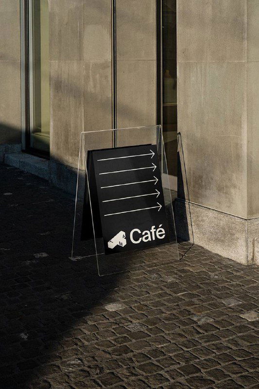 Уличный указатель‑«A‑frame» с чёрным фоном и стрелками навигации; демонстрирует применение фирменной графики и ориентиры для посетителей музея.