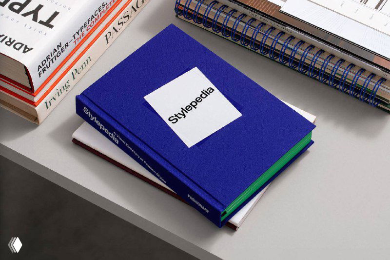 Книга «Stylepedia» лежит на столе рядом с другими изданиями; видно текстильную обложку синего цвета и контрастные зелёные торцы страниц.