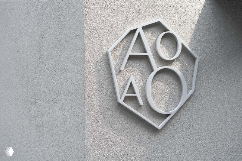 Металлический объёмный логотип AOAO на фактурной стене в светлом варианте, сочетание благородного зелёного и нейтральных серых тонов в брендинге.