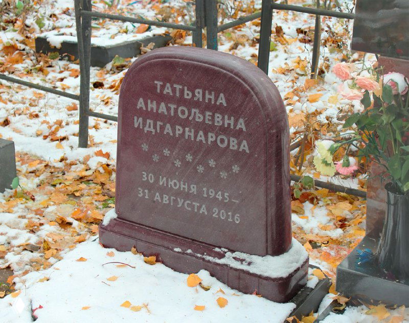 Надгробие на открытом участке кладбища среди опавших листьев и лёгкого снега, тёмный камень и буквы, общий план демонстрирует контекст.