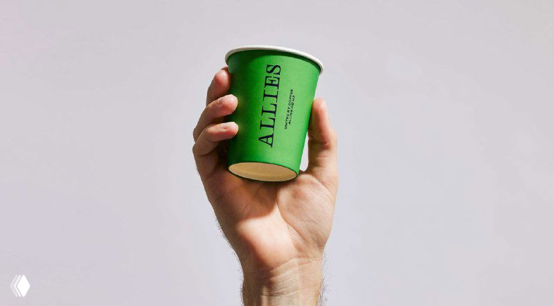 Крупный план руки, держащей маленький зелёный бумажный стаканчик ALLIES на нейтральном фоне — акцент на тактильности и логотипе продукта