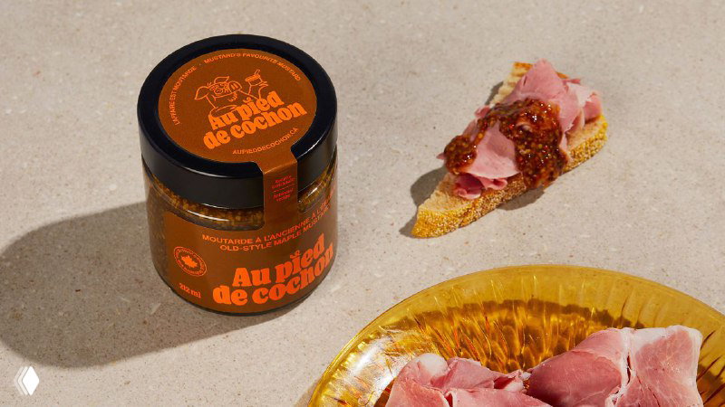 Банка с продуктом и ломтик хлеба с ветчиной на столе: оформление баночки в приглушённых тонах с ярким оранжевым логотипом Au Pied de Cochon.