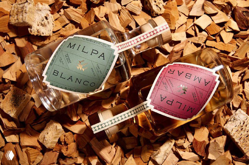 Две бутылки Milpa лежат на древесных щепках: бирки зелёного и розового цветов крупным планом, подчёркнута текстура упаковки и материал.