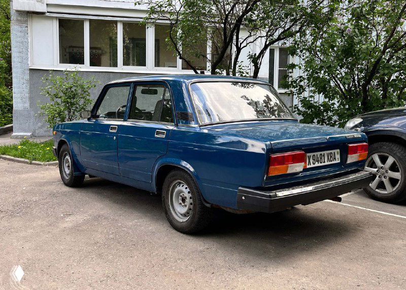 Синий классический седан припаркован во дворе: на корме видно российский регистрационный знак, городской фон и тротуар.